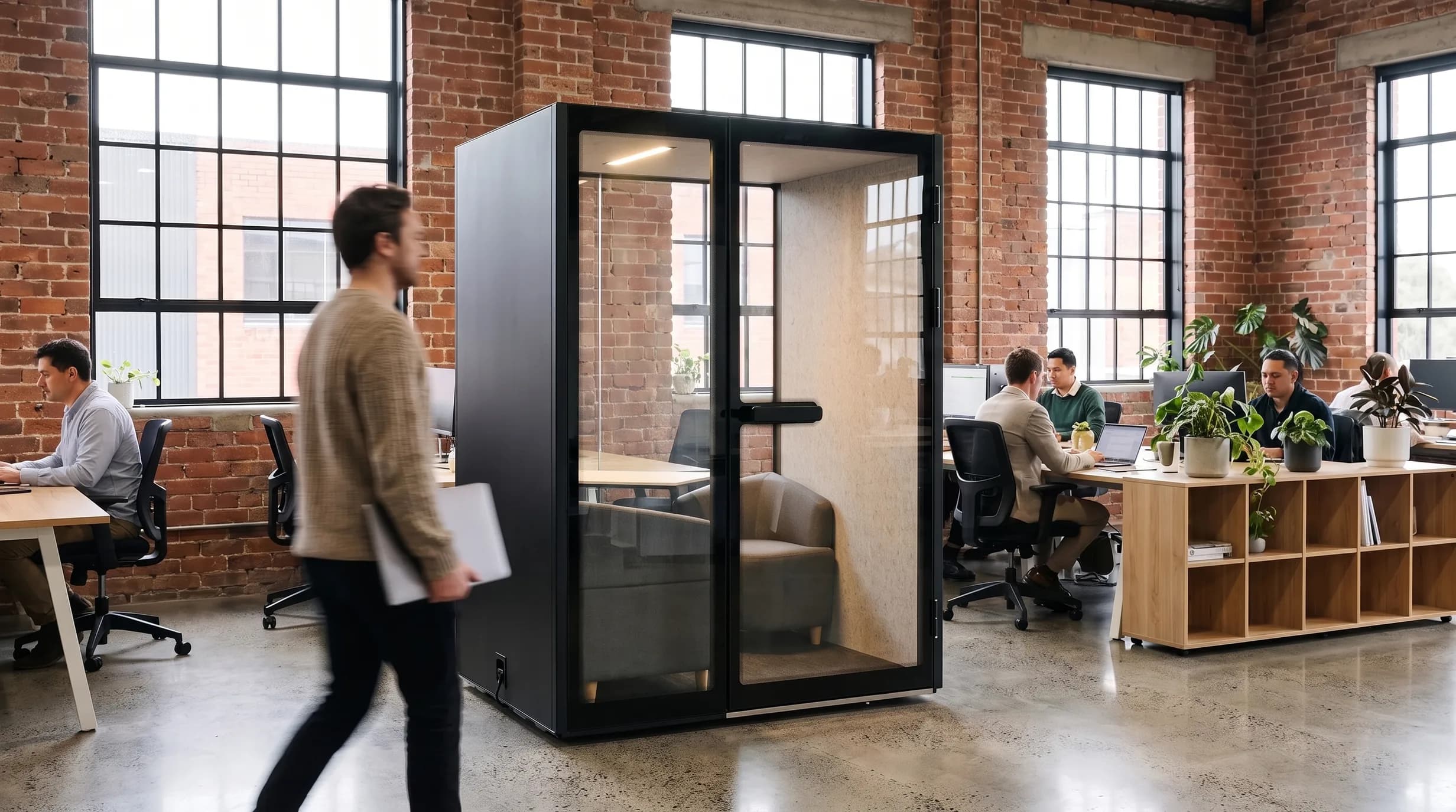 M2-Pod meeting pod in a brick-warehouse open-plan office, a person walking past with a laptop