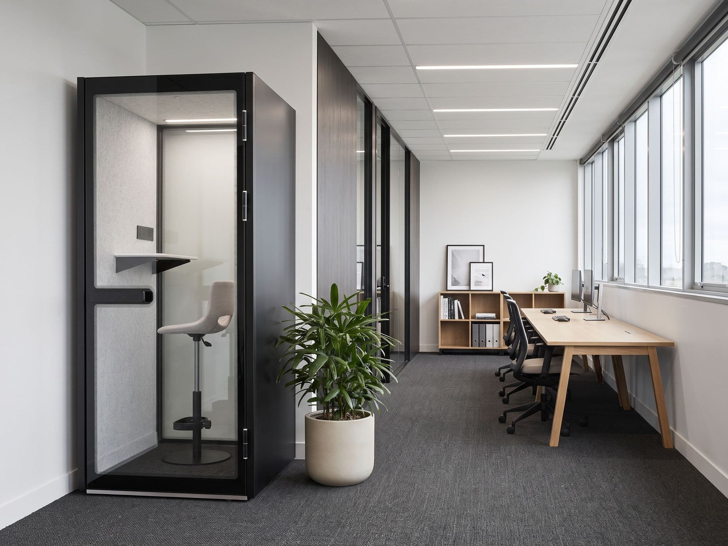 An S-Pod phone booth installed against a white wall in a quiet office, with a potted plant in the foreground and desks along the windows beyond