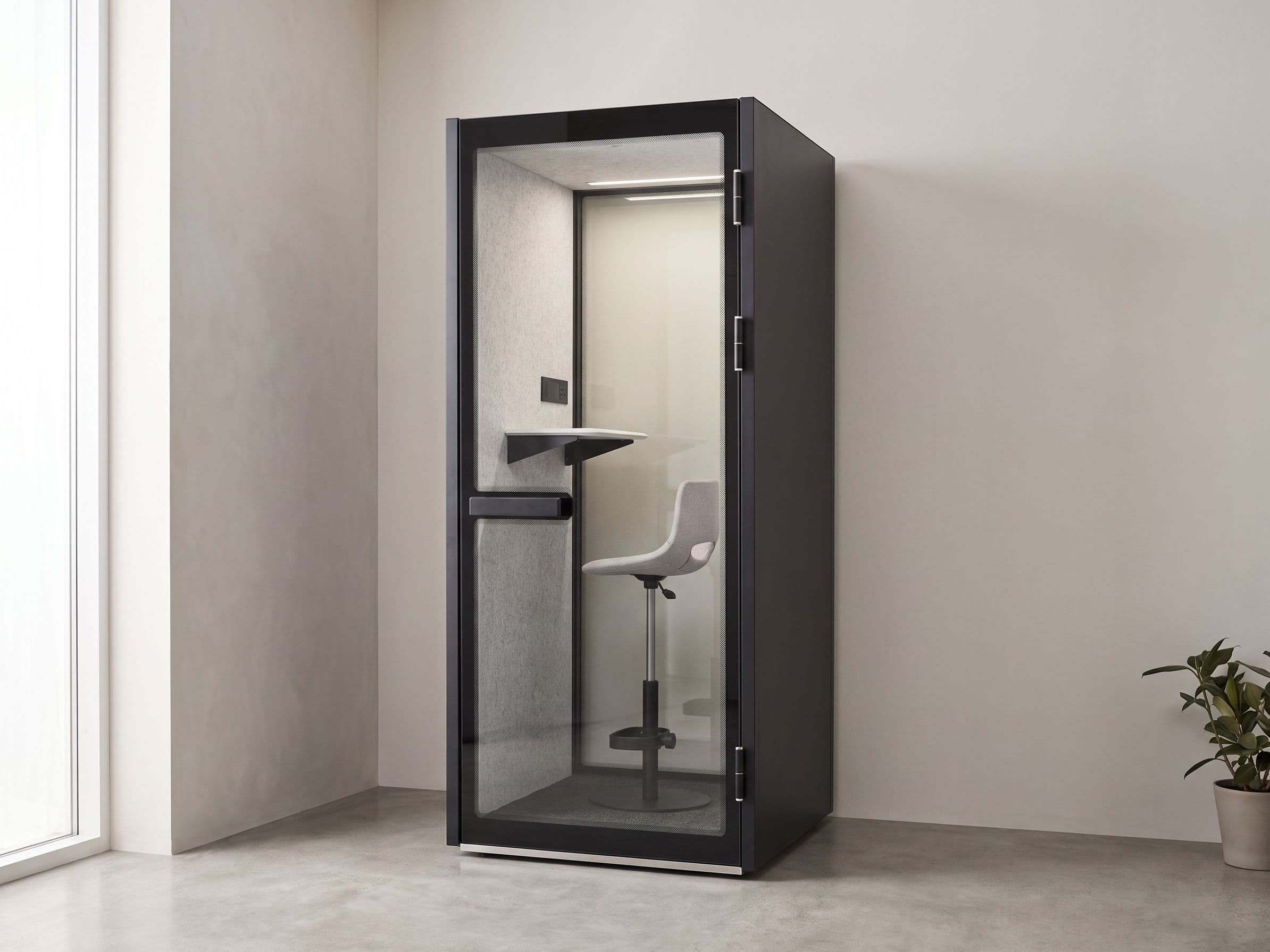 An S-Pod phone booth installed in the corner of a quiet, uncluttered interior, lit by soft daylight from a window to one side