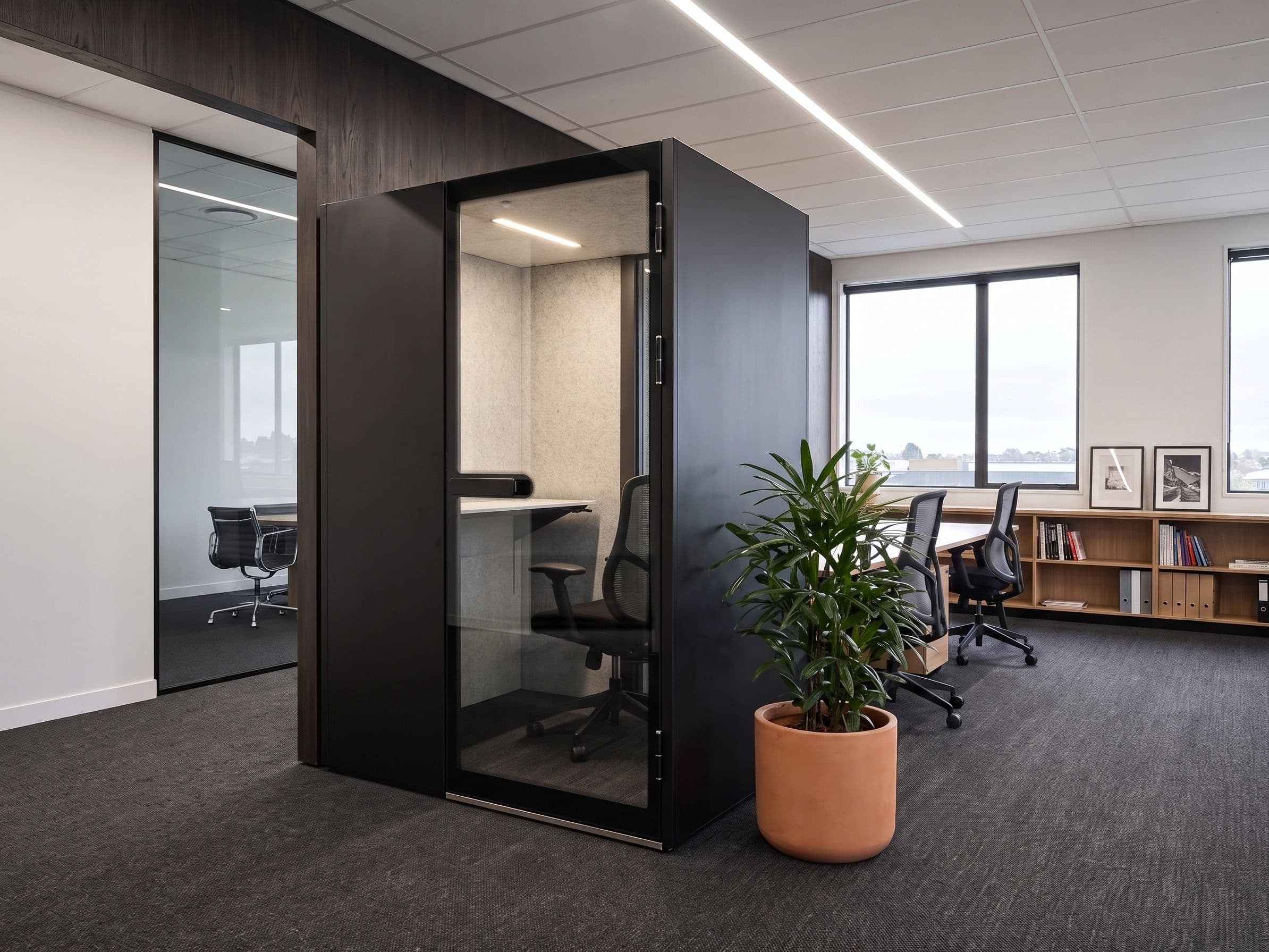 An MW-Pod installed against a dark timber wall in a quiet office, with a potted plant in a terracotta pot in the foreground and desks along the windows beyond
