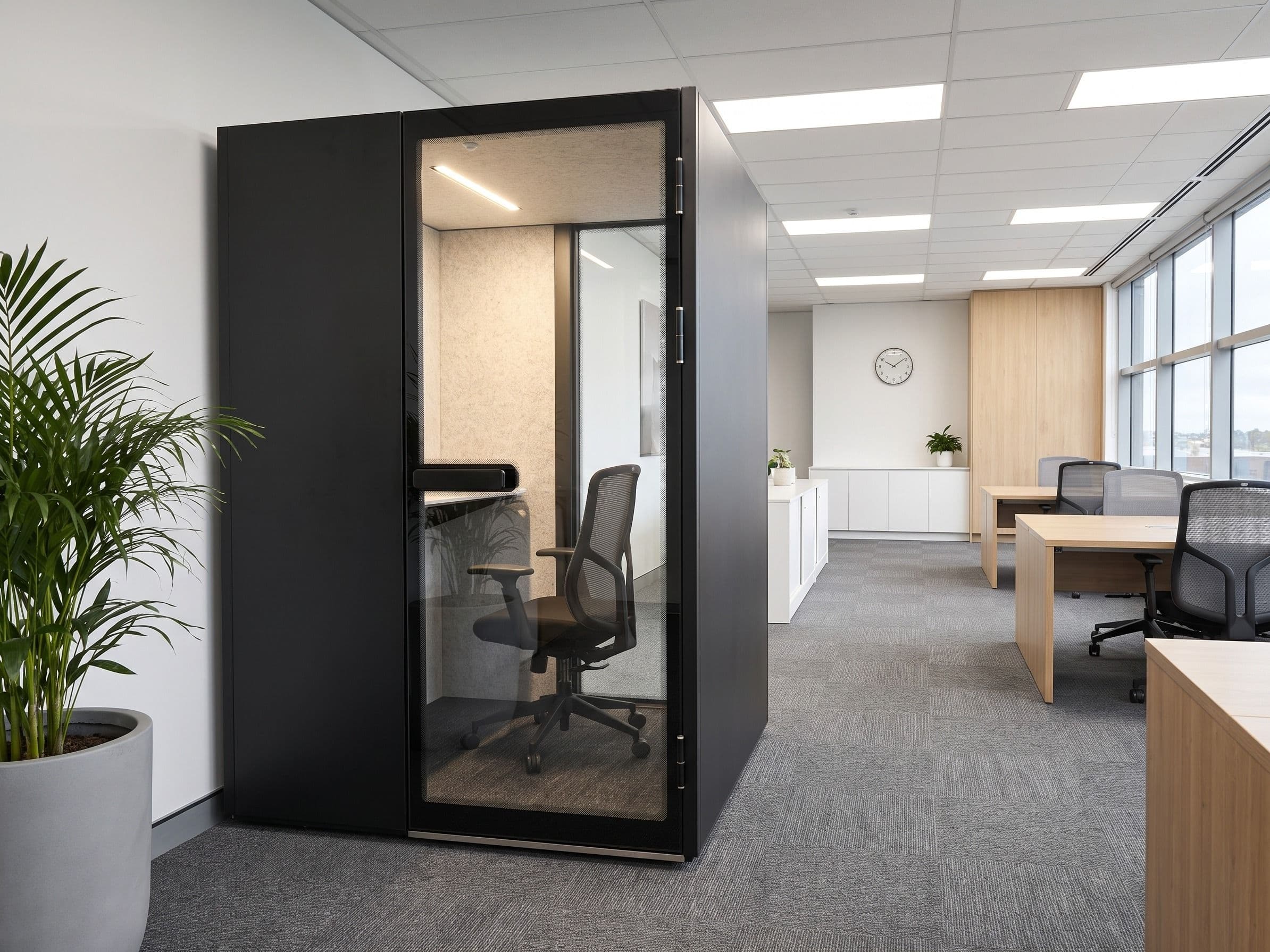 An MW-Pod installed against a white wall in a tidy open-plan office, with oak desks, a low white credenza and a wall clock beyond, and a potted palm in the foreground
